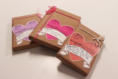 Three books wrapped with brown paper and a bow have a pink or purple-colored heart on the center with each book's genre, barcode, and their first line. 